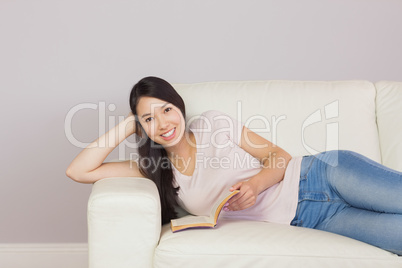 Happy asian girl lying on the sofa reading book smiling at camer