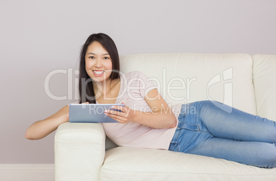 Pretty asian girl lying on the sofa using digital tablet smiling