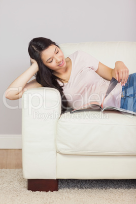 Cheerful asian girl lying on the couch reading a magazine