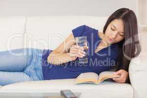 Happy asian girl lying on the sofa reading a novel and drinking