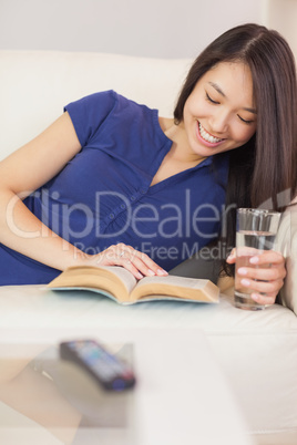 Smiling asian girl lying on the sofa reading a novel and drinkin