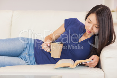 Beautiful asian girl lying on the sofa reading a novel and drink