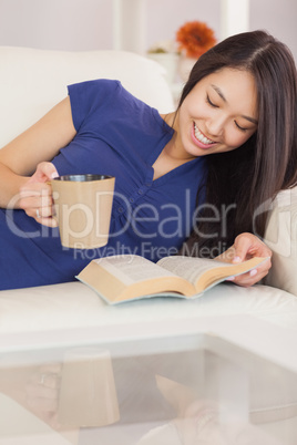 Happy asian girl lying on the sofa reading a novel and drinking