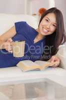 Smiling asian girl lying on the sofa reading a novel and drinkin