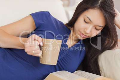 Attractive asian girl lying on the sofa reading a novel and drin