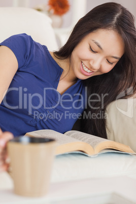 Relaxed asian girl lying on the sofa reading a novel and drinkin