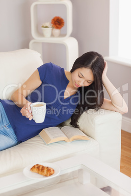 Pretty young asian woman lying on the sofa reading a book holdin