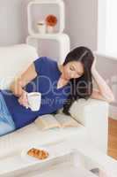 Pretty young asian woman lying on the sofa reading a book holdin
