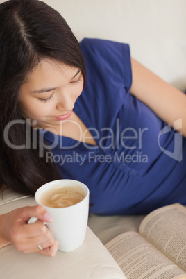 Young asian woman lying on the sofa reading a book holding her c