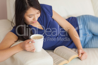 Happy young asian woman lying on the sofa reading a book holding