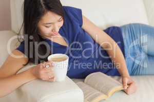 Happy young asian woman lying on the sofa reading a book holding