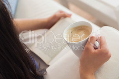 Woman lying on the sofa reading a book holding her coffee