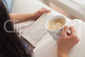 Woman lying on the sofa reading a book holding her coffee