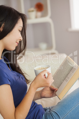 Young asian woman sitting on the sofa reading a book holding her