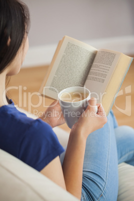 Young woman sitting on the sofa reading a book holding her coffe