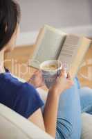 Young woman sitting on the sofa reading a book holding her coffe
