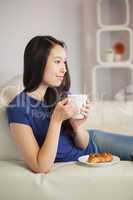 Young asian woman sitting on the sofa having coffee with a pastr