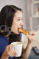 Young asian woman sitting on the sofa having coffee and eating a