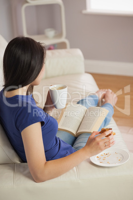 Young woman reading a book and eating pastry with coffee