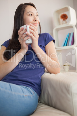 Thoughtful young asian woman sitting on the couch holding mug