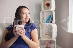 Thinking young asian woman sitting on the couch holding mug