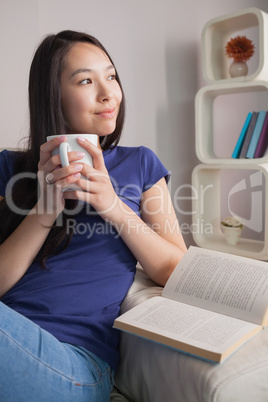 Thinking asian woman sitting on the couch holding mug of coffee
