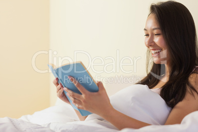 Smiling young asian woman sitting in bed reading a book