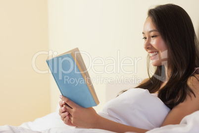 Happy young asian woman sitting in bed reading a book