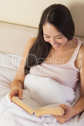 Cheerful young asian woman sitting in bed reading a book