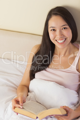 Cheerful young asian woman sitting in bed reading a book looking