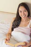 Cheerful young asian woman sitting in bed reading a book looking