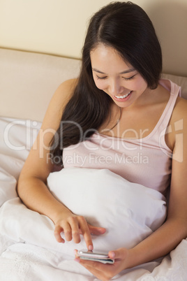 Cheerful young asian woman sitting in bed texting on her smartph