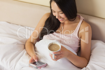 Cheerful young asian woman sitting in bed texting on her smartph
