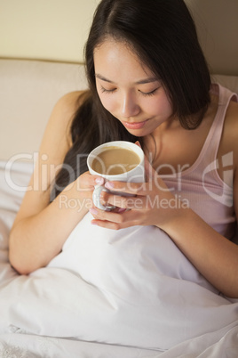 Cheerful young asian woman sitting in bed smelling her morning c