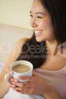 Cheerful young asian woman sitting in bed holding her morning co