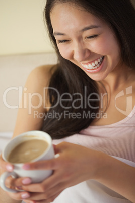Laughing young asian woman sitting in bed holding her morning co