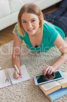 Cheerful young woman lying on floor using tablet to do her assig