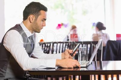 Serious businessman using phone while working on laptop