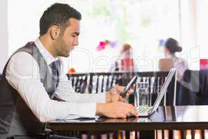 Serious businessman using phone while working on laptop