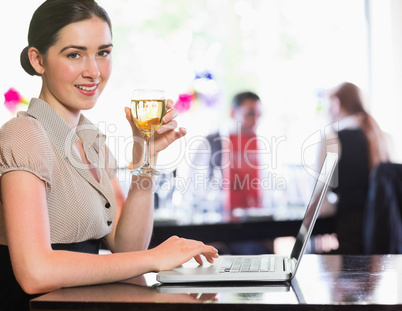 Happy businesswoman holding wine glass using laptop and looking