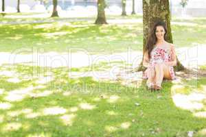Stylish cheerful brunette sitting and leaning against tree