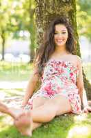 Stylish smiling brunette sitting and leaning against tree