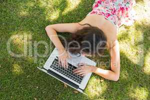 Stylish brunette lying on the grass typing at her laptop