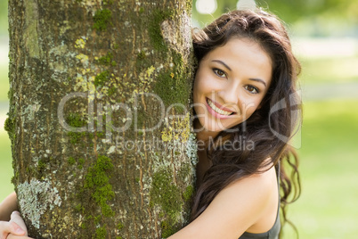 Casual smiling brunette embracing a tree looking at camera