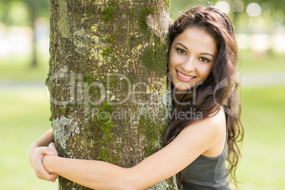 Casual cheerful brunette embracing a tree looking at camera