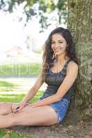 Casual cheerful brunette sitting leaning against tree