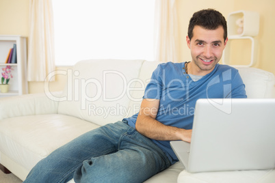 Casual smiling man sitting on couch using laptop looking at came