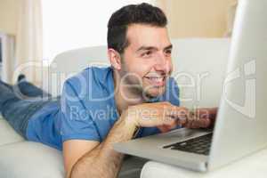 Casual cheerful man sitting on couch using and looking at laptop