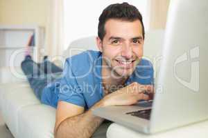 Casual cheerful man sitting on couch using laptop looking at cam