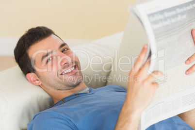 Casual smiling man lying on couch holding newspaper